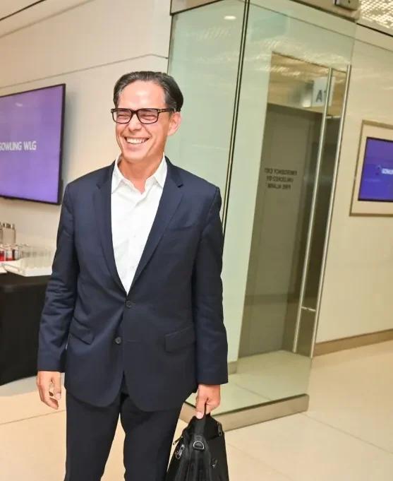 David Legendre, three-quarter view, smiling, in a navy suit, white open-collar shirt, and glasses, walking through the Gowling WLG lobby with a bag. A professional gathering — not a speaking appearance.